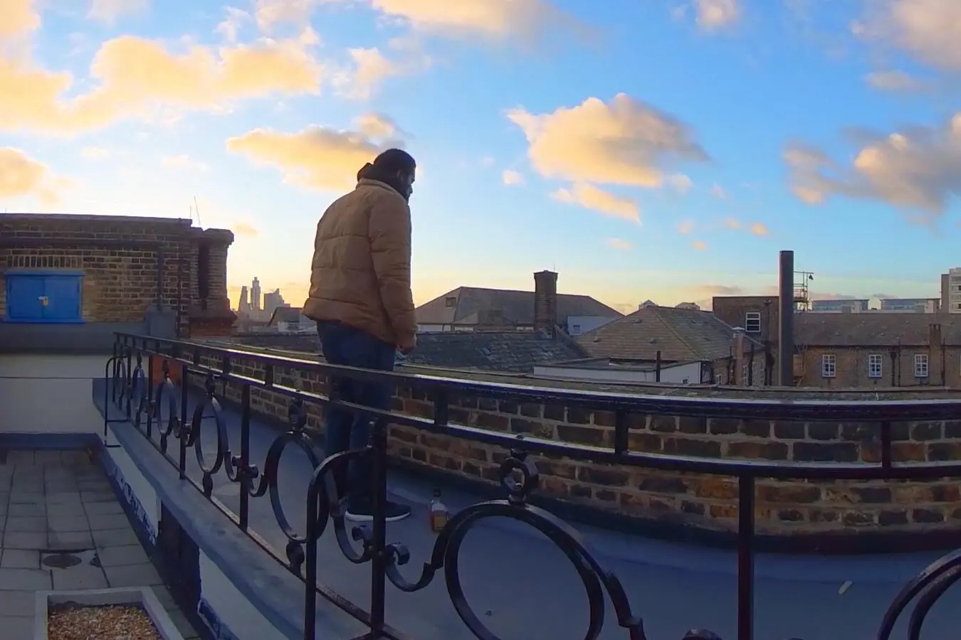 The rooftop scene: a man in a padded jacket stands alone on a flat roof under a low sun, looking down